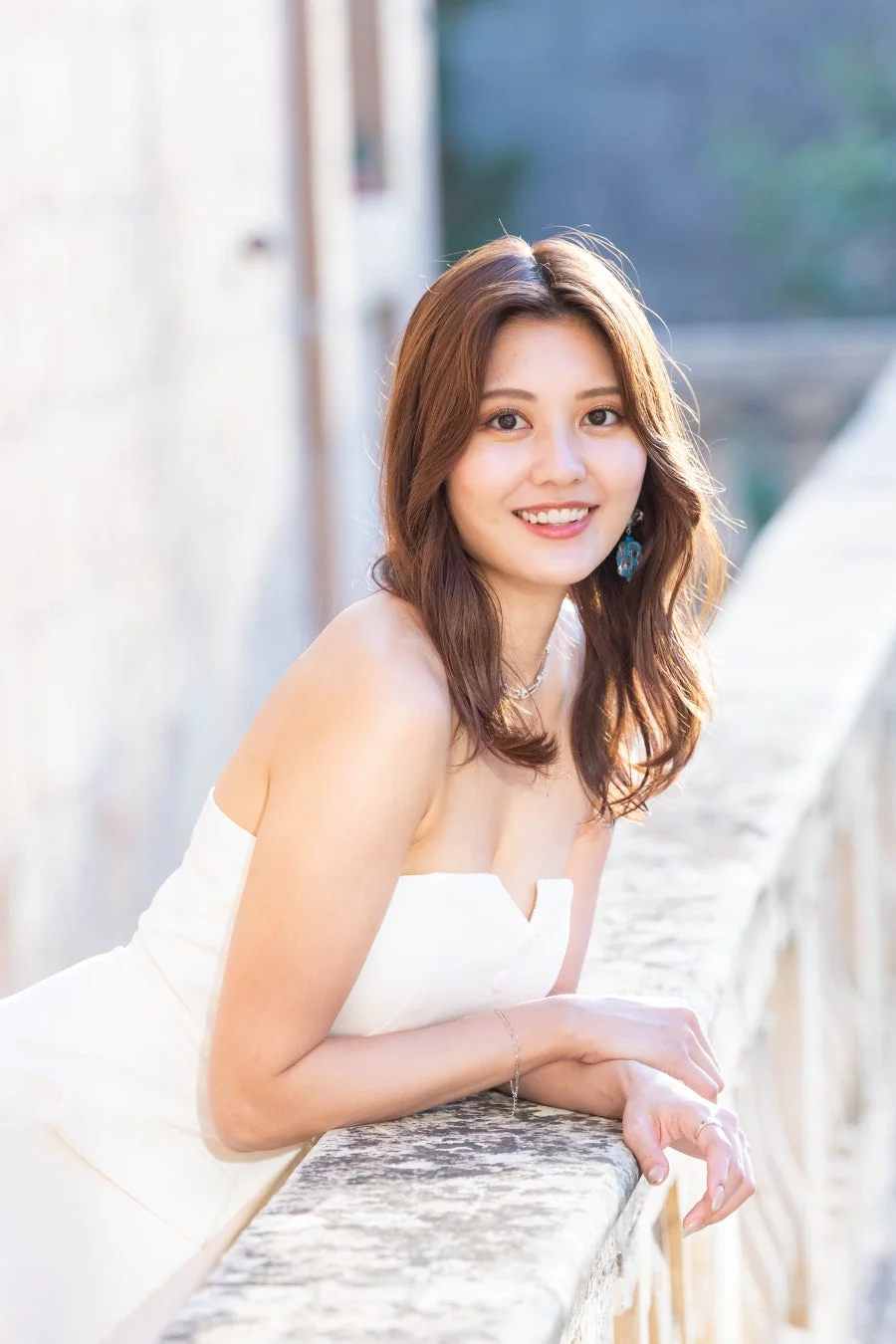 白いストラップレスドレスを着た笑顔の若いアジア人女性が、屋外で石の手すりに寄りかかっているポートレート。柔らかな日差しが彼女の髪を照らし、明るく美しい雰囲気を醸し出しています。