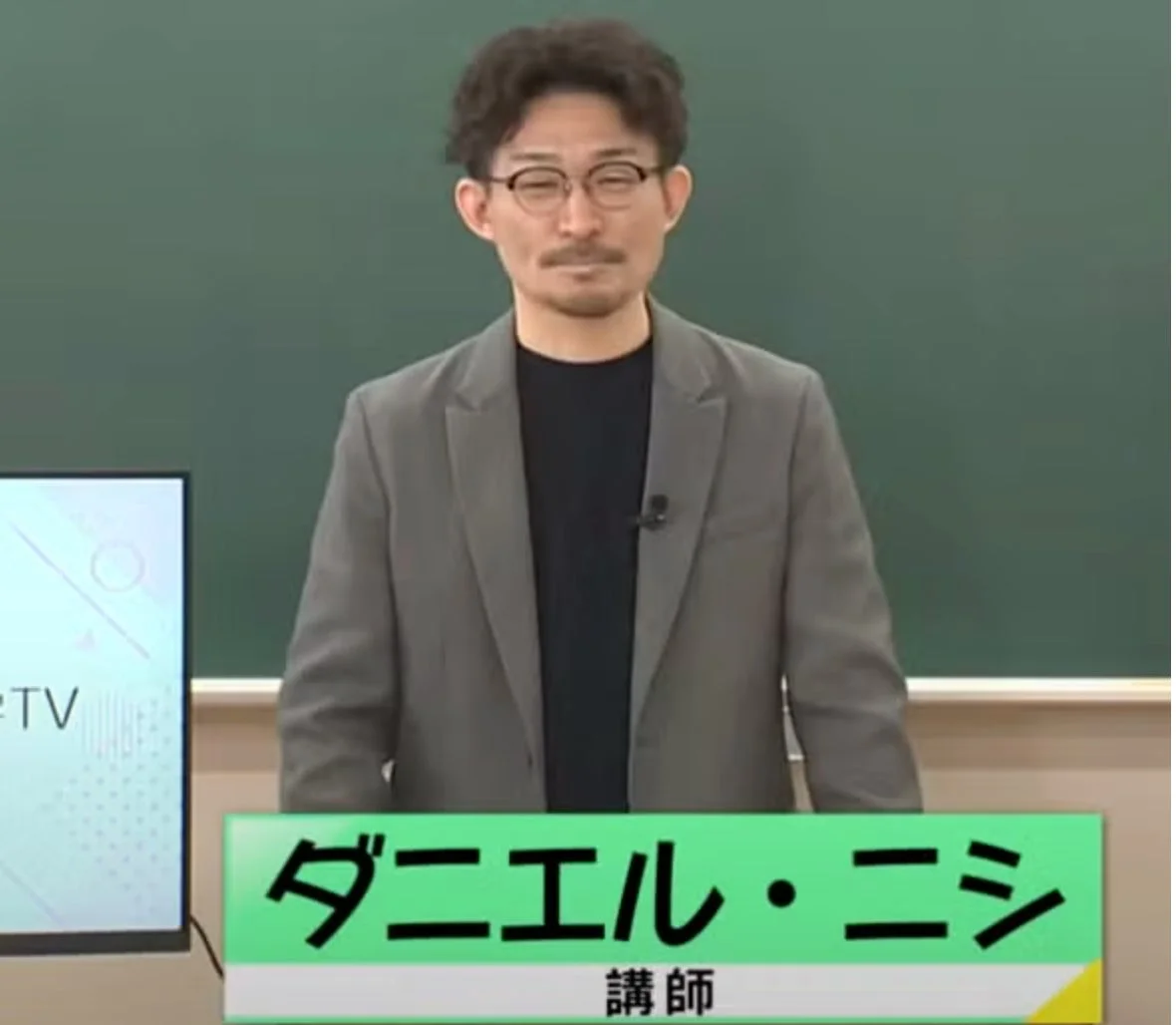 緑色の黒板を背景に、眼鏡をかけた男性講師「ダニエル・ニシ」が立っている画像です。