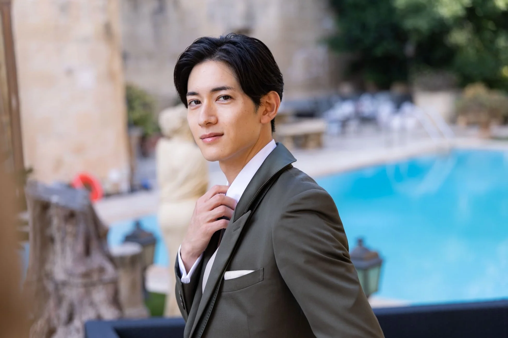 屋外のプールサイドで、若く端正な顔立ちの男性がカメラに向かって微笑んでいます。ダークグリーンのスーツを着ており、手で襟元を整える仕草をしています。
