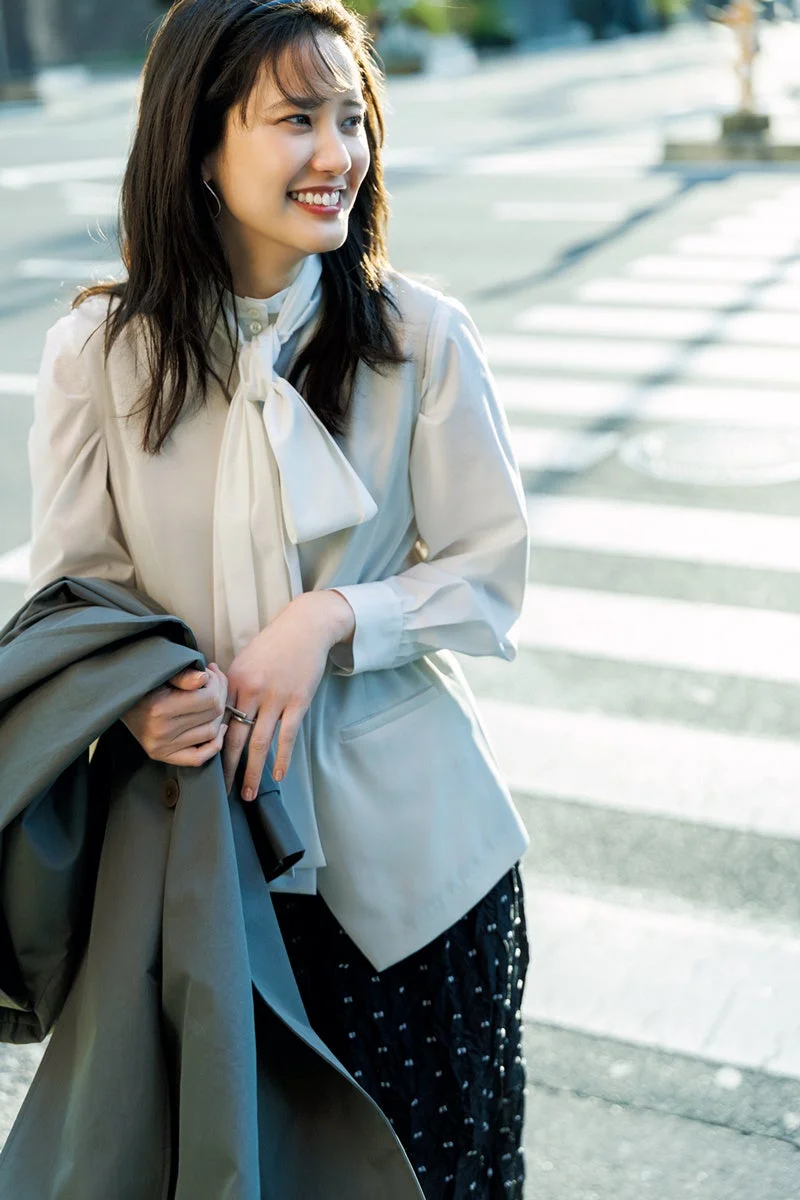 太陽の光が当たる屋外で、白いボウタイブラウスと黒いスカートを着用し、笑顔で振り返る若い女性。手にグレーのジャケットを持ち、明るい表情で街角に佇んでいます。
