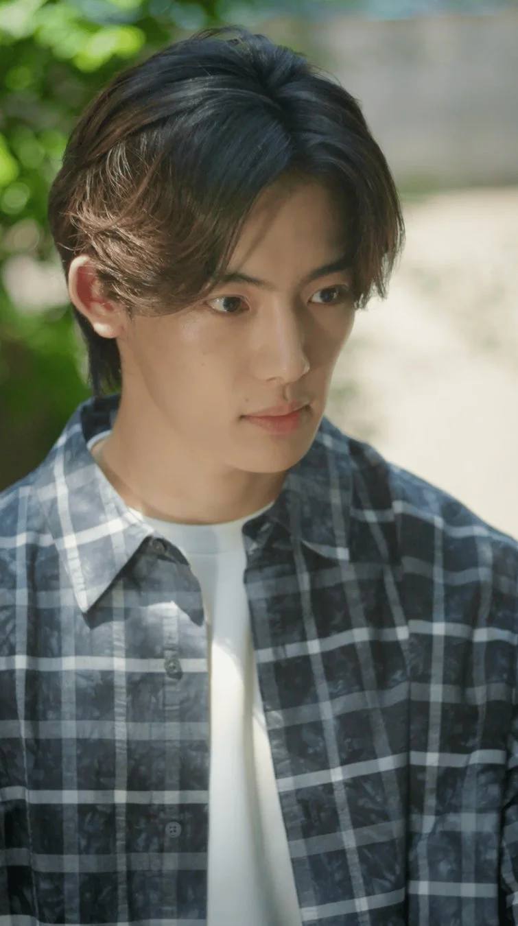 若い男性が屋外でチェック柄のシャツを羽織り、白いTシャツを着ています。少し下を向いた表情で、背景には緑豊かな自然がぼかされています。