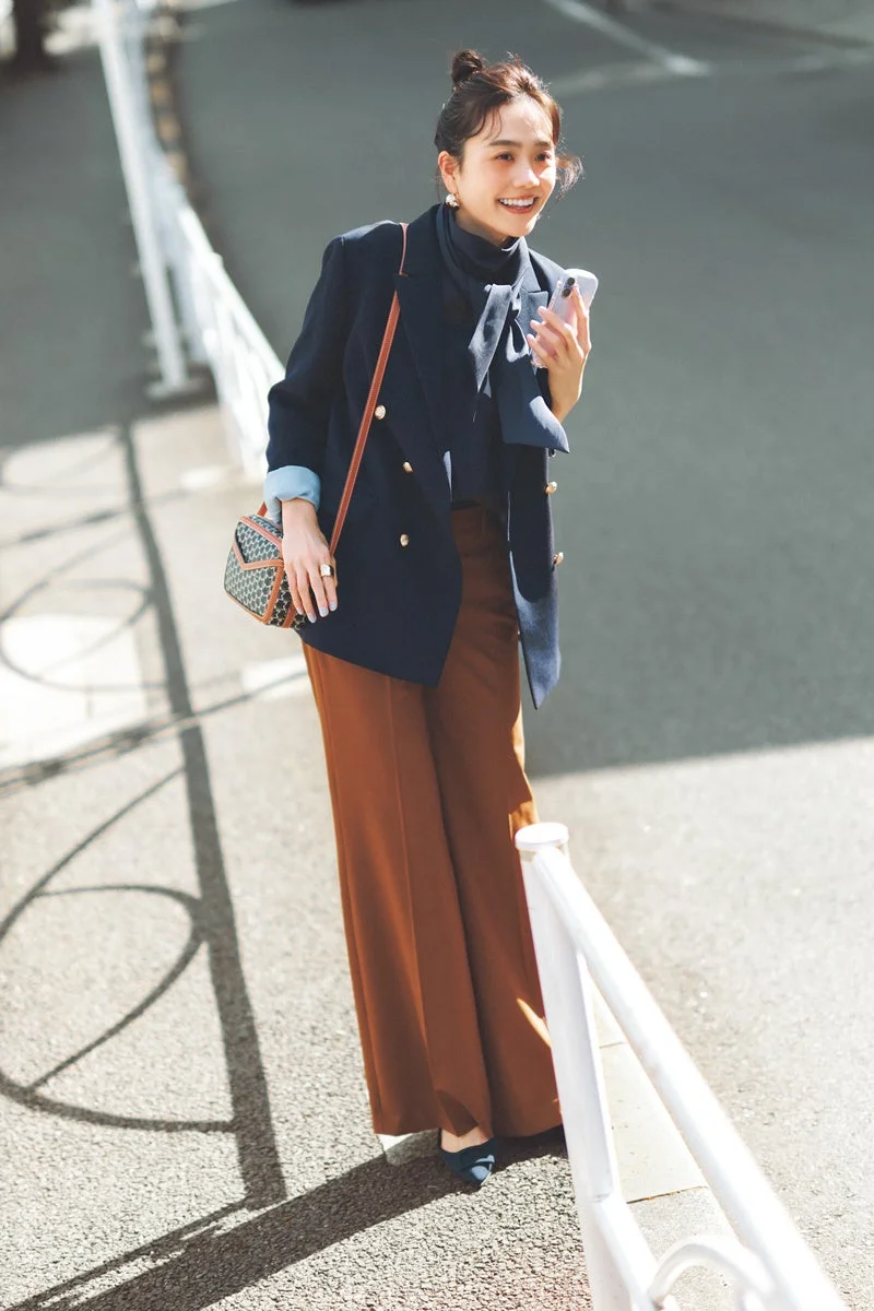 ネイビーのブレザーとブラウンのワイドパンツを着用し、笑顔で歩く女性の全身ショットです。スマートフォンを持ち、上品なショルダーバッグを肩にかけています。日中のストリートで、光と影のコントラストが印象的です。