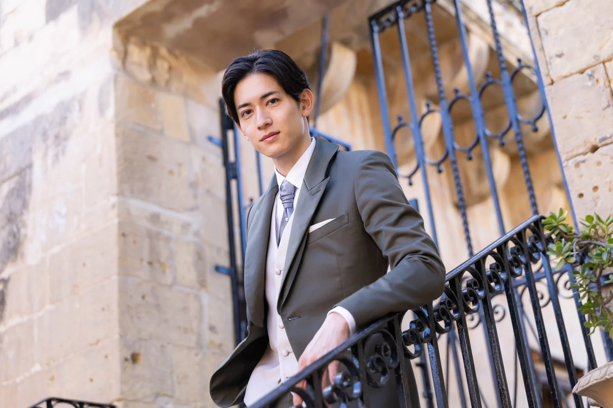 オリーブグリーンのスーツを着用した若いアジア人男性が、歴史的な石造りの建物と装飾的な鉄柵を背景に立つポートレート