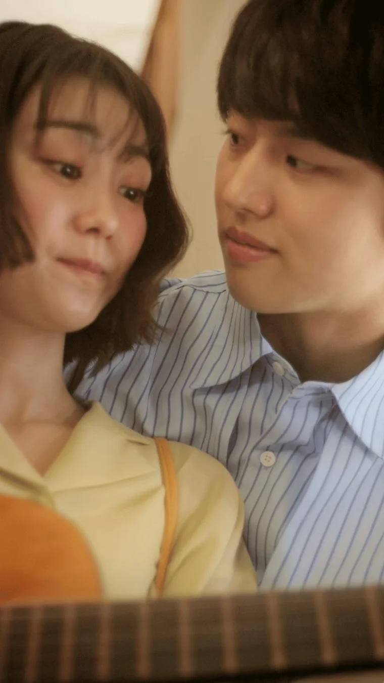 若い男女が寄り添っているクローズアップ写真です。女性はギターを抱えているようで、二人の間に穏やかで親密な雰囲気が漂っています。