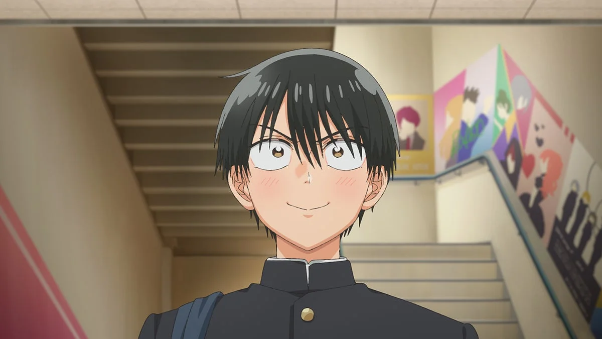 アニメ調の黒髪の男子学生が、学校の階段で制服姿で笑顔を見せている。頬は赤く染まり、少し照れているような表情をしている。背景にはカラフルなポスターが飾られている。