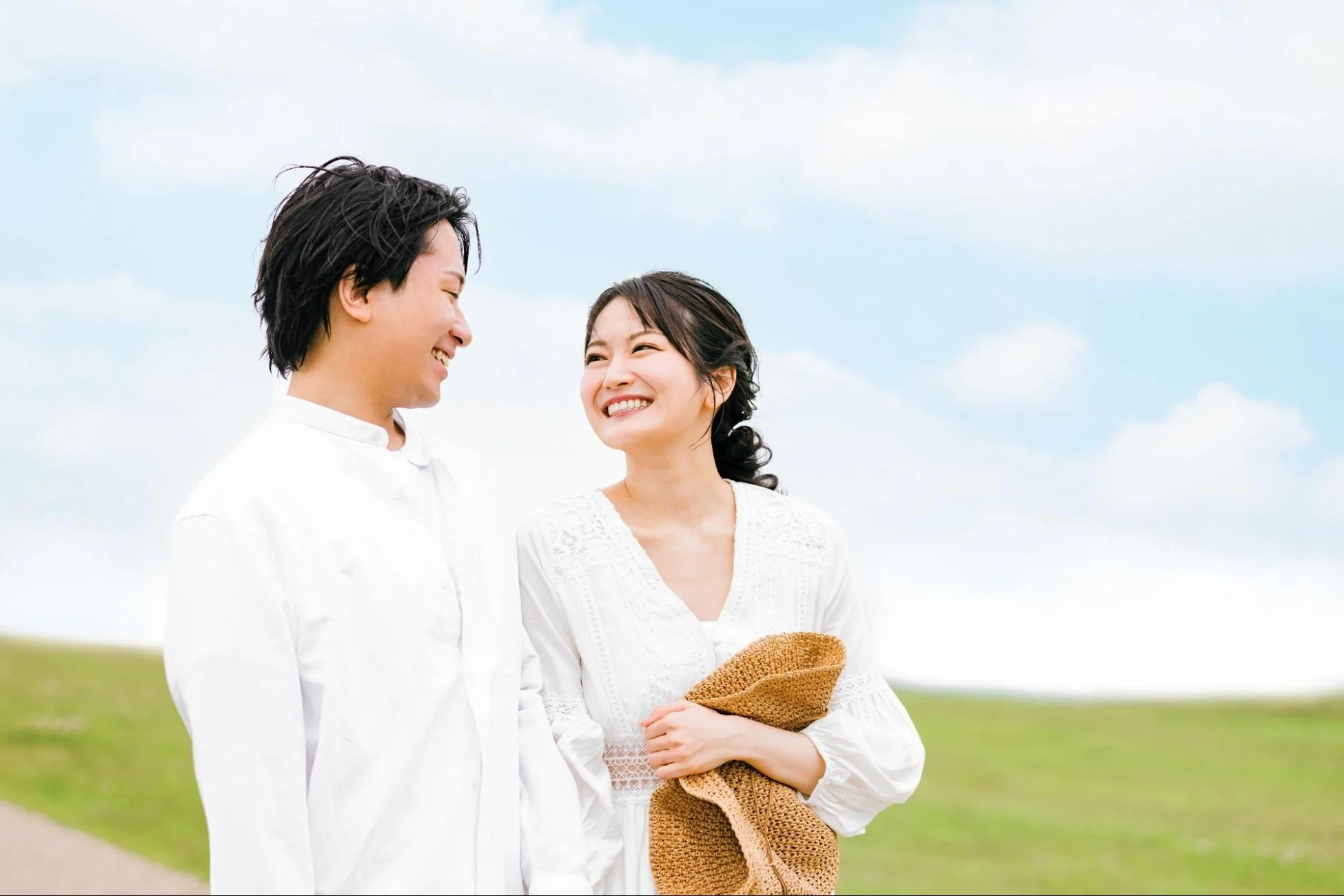 青空と緑の草原の下、白い服を着た若いカップルが互いに笑顔を向け合い、幸せそうに散歩している様子