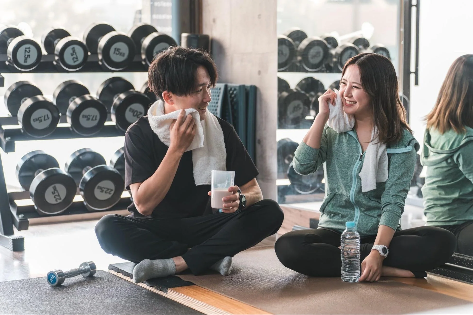 ジムでトレーニング後に休憩している男女の様子。男性はプロテインを、女性は水を飲み、タオルで汗を拭きながら笑顔で会話している。背景には様々な重さのダンベルが並んでいる。