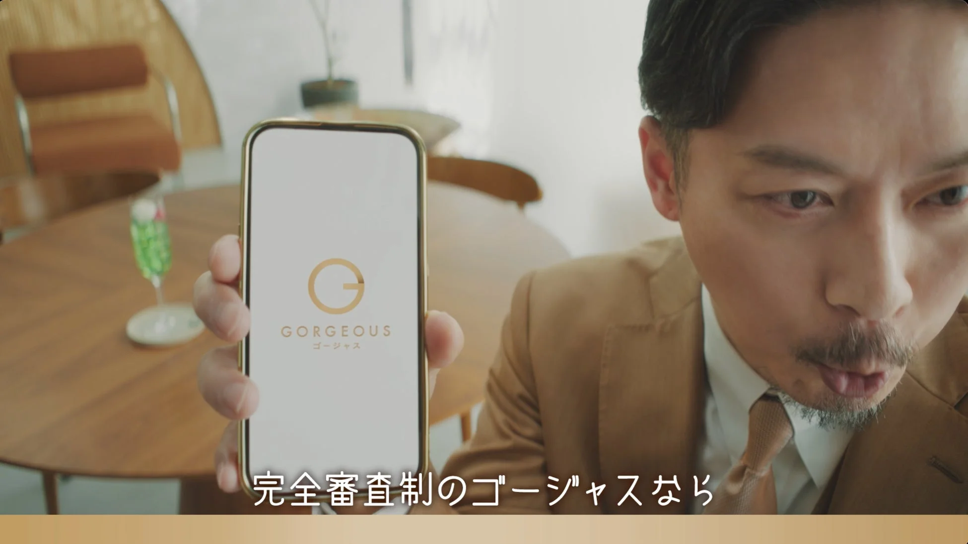 スーツ姿の男性がスマートフォンで「ゴージャス」アプリの画面を見る場面