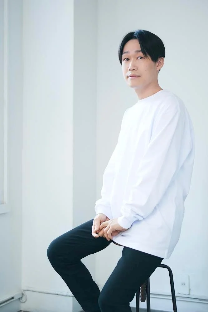 白い長袖シャツと黒いパンツを着用し椅子に座って微笑む大塚陽介氏