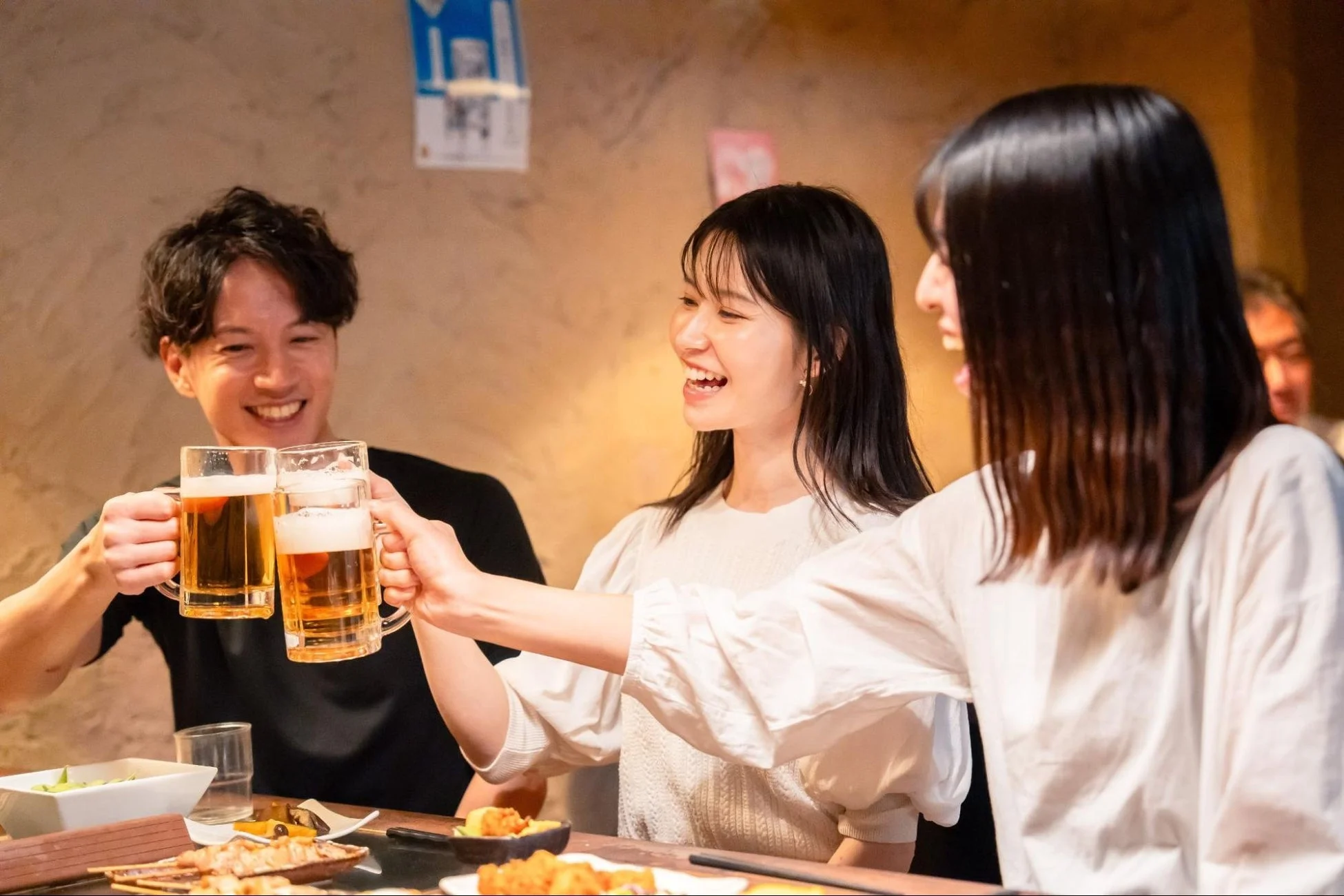 友人同士と思われる3人が居酒屋でビールジョッキを手に乾杯している様子です。皆笑顔で、楽しい時間を過ごしていることが伝わります。テーブルには料理も並んでいます。