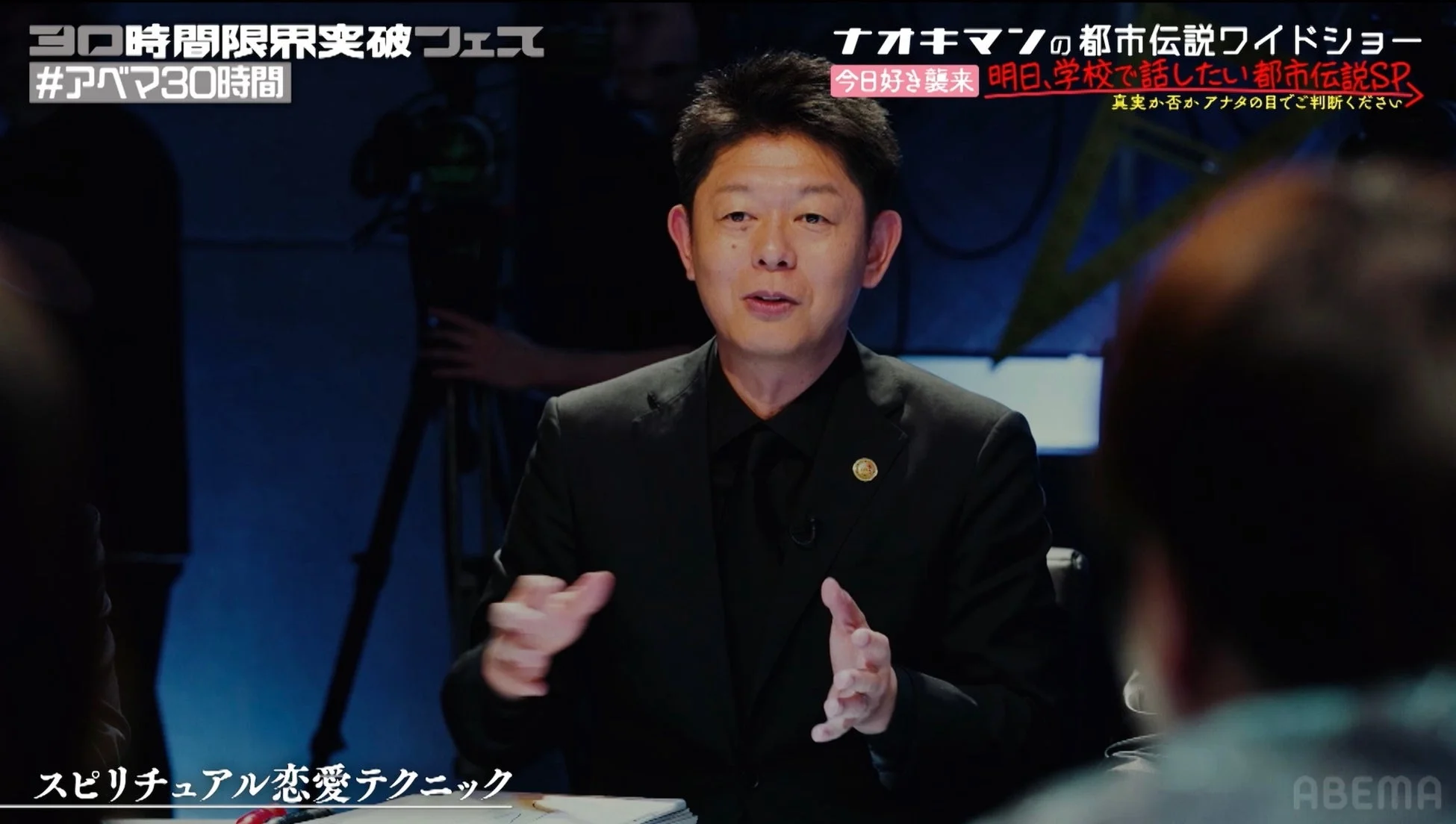 「ナオキマンの都市伝説ワイドショー」に登場する男性が、スタジオで熱心に話している場面。番組は「30時間限界突破フェス」の一部で、スピリチュアルな恋愛テクニックや都市伝説がテーマのようだ。