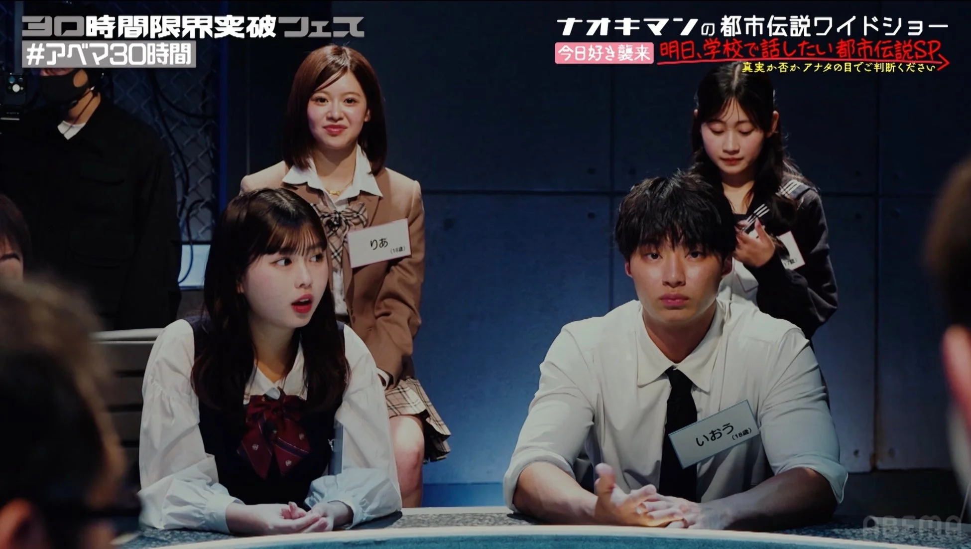 ABEMAの『30時間限界突破フェス』内で放送された『ナオキマンの都市伝説ワイドショー』の一場面。若者たちが集まり、真剣な表情で都市伝説について討論している様子が伺える。番組は『明日、学校で話したい都市伝説SP』と題されており、りあといおうという18歳の出演者の名札が見える。