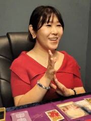 赤い服を着たアジア人女性が笑顔で拍手している様子です。テーブルにはタロットカードのようなものが並べられており、占い師か鑑定を受けている場面を思わせます。