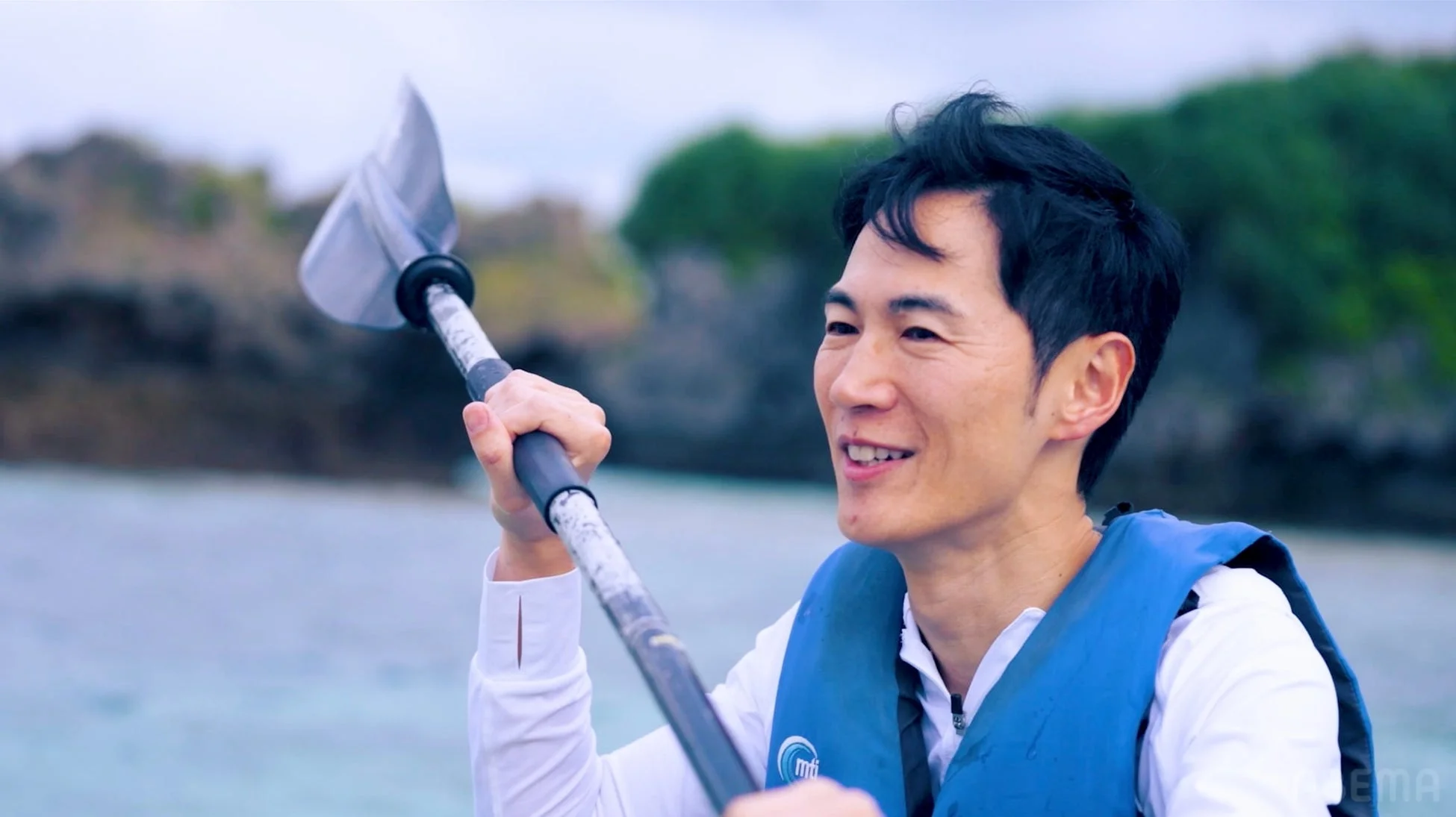 青いライフジャケットを着た男性がカヤックのパドルを持ち、水上で笑顔を見せています